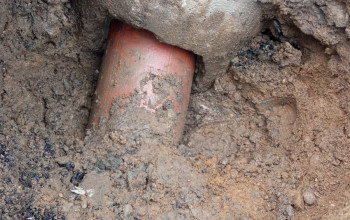 To zdjęcie przedstawia pomarańczową rurę kanalizacyjną częściowo zakopaną w błotnistym wykopie ziemnym. Nad rurą widoczny jest duży, betonowy blok, który stabilizuje instalację.