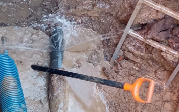 To zdjęcie przedstawia wykop ziemny z awarią, z którego tryska woda z uszkodzonego czarnego rurociągu. Na pierwszym planie widoczna jest łopata, a obok niebieska karbowana rura osłonowa.