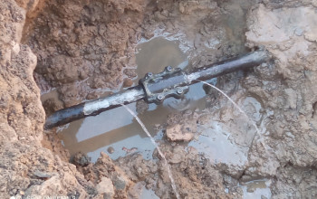 Zdjęcie przedstawia grubą, czarną rurę częściowo zanurzoną w błotnistej wodzie, wewnątrz wykopu ziemnego. Na środku rury widoczny jest metalowy pierścień zaciskowy, z którego tryska woda.