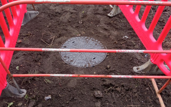 Okrągła, metalowa pokrywa studni rewizyjnej znajduje się pośrodku zabłoconego wykopu.
Czerwone plastikowe barierki otaczają teren prac.
