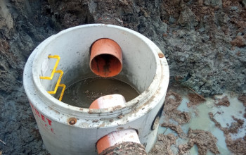 Betonowa studnia z pomarańczowymi rurami znajduje się w głębokim, błotnistym wykopie.
Widoczne są żółte metalowe stopnie wewnątrz studni, co wskazuje na prace przy budowie sieci wod-kan.