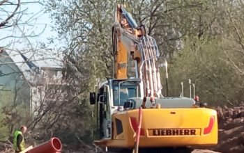 Duża, żółta koparka stoi na błotnistym terenie budowy, otoczonym zielonymi drzewami. Na pierwszym planie widoczne są stosy pomarańczowych rur kanalizacyjnych, a obok stoi pracownik.
