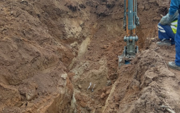 Duży, głęboki wykop w ziemi ukazuje brązowe, ubite warstwy gleby, powstałe podczas prac koparką. W dole wykopu widoczny jest fragment czerwonej rury, a nad nim ramię i łyżka maszyny.