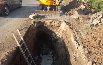 Duży, głęboki wykop w ziemi, wypełniony błotem i wodą, odsłania miejsce awarii przy drodze.
Obok wykopu stoi żółta koparka.